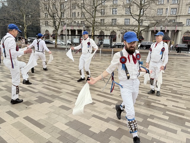Newtowne Morris performing Fieldtown Dearest Dickie on Copley Plaza, two lines of dancers in whites, suspenders, and blue caps facing each other with two of the dancers facing out of the set.