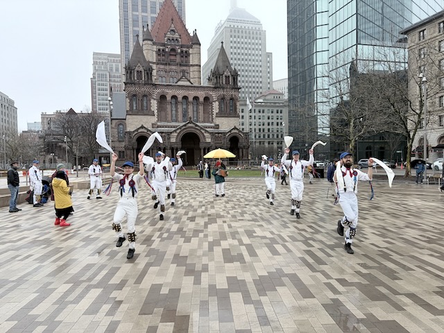 Newtowne Morris performing Ducklington The Fox on Copley Plaza across from Trinity Church, two lines of dancers in whites, suspenders, and blue caps fancing the camera and waving their hankies.