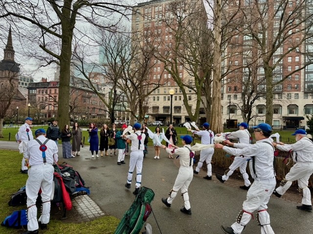 Newtowne Morris performing Stanton Harcourt Nutting Girl on a path in the Public Garden across from the Newbury Hotel, two lines of dancers in whites, suspenders, and blue caps facing away from the camera and flicking their hankies forward towards the musican and audience.