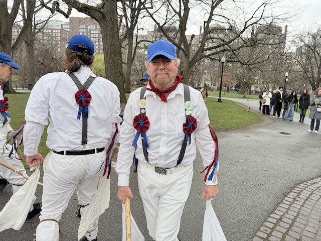 Newtowne Morris performing Stanton Harcourt Greensleaves on a path in the Public Garden, two dancers in whites, suspenders, and blue caps holding sticks and hankies, one facing away and the other facing towards the camera as they pass in the hey.