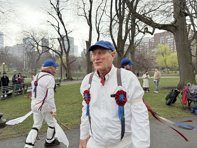 Newtowne Morris performing Stanton Harcourt Greensleaves on a path in the Public Garden near the pond, two dancers in whites, suspenders, and blue caps holding sticks and hankies, one facing to the right and the other to the left.