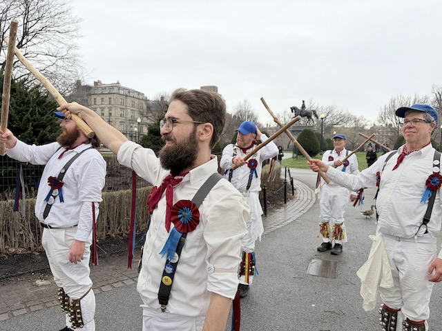 Newtowne Morris performing Fieldtown Balance the Straw on a path in the Public Garden near the equestrian statue of George Washington, two lines of dancers in whites, suspenders, and blue caps holding sticks across the set.