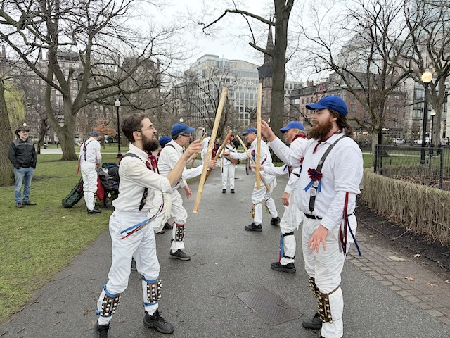 Newtowne Morris performing Fieldtown Balance the Straw on a path in the Public Garden, two lines of dancers in whites, suspenders, and blue caps facing across the set and tossing sticks to each other, the musician at the other end of the set.