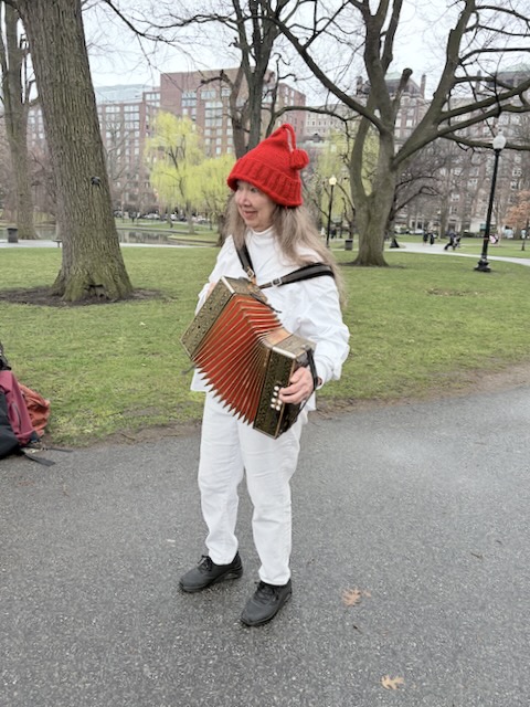 Newtowne Morris musician in whites and a bright red knit cap standing with a melodeon a path in the Public Garden.