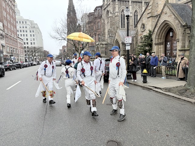 Newtowne Morris performing Stanton Harcourt Nightingale on Newbury Street in front of Emmanuel Church, dancers in whites, suspenders, and blue caps holding sticks to their sides and moving in different directions in mirror heys for three.