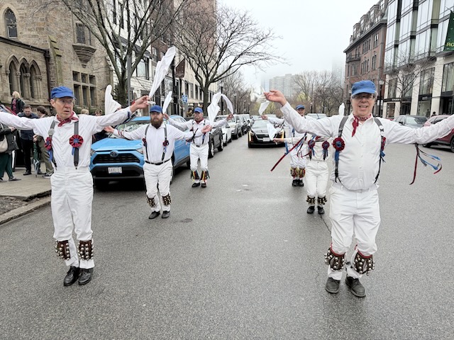 Newtowne Morris performing Fieldtown Shepherd's Hey on Newbury Street in front of Emmanuel Church, two lines of dancers in whites, suspenders, and blue caps facing the camera and holding their hands out with hankies.