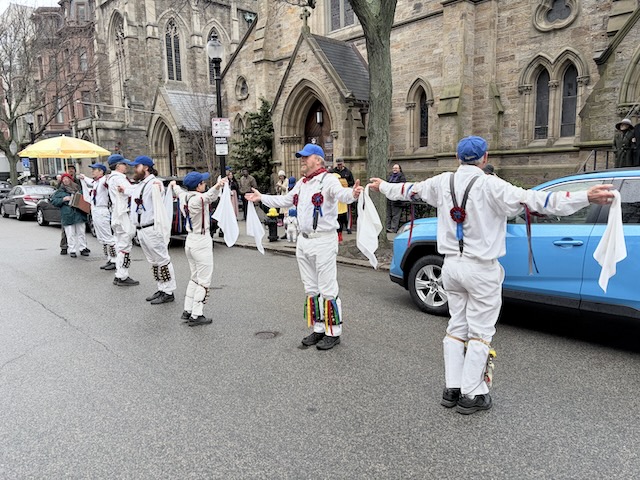 Newtowne Morris performing Fieldtown Shepherd's Hey on Newbury Street in front of Emmanuel Church, a single line of dancers in whites, suspenders, and blue caps facing alternating directions with their hands out holding hankies.